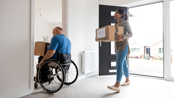 A man in a wheelchair and his companion carry boxes into an empty house.