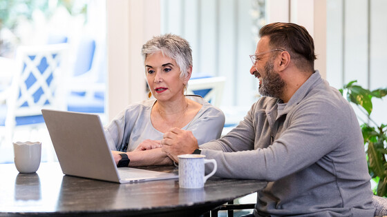 A woman uses her laptop at a table, while seated next to a bespectacled man who is smiling at her.