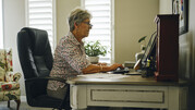 A woman with gray hair and glasses sits at a desk with a computer in a home office setting.