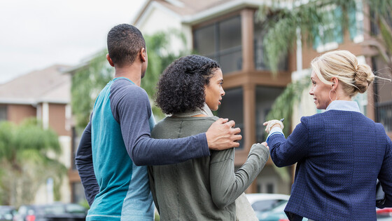 A leasing agent speaks to a woman and man about a rental property, while pointing to the dwelling across the street.