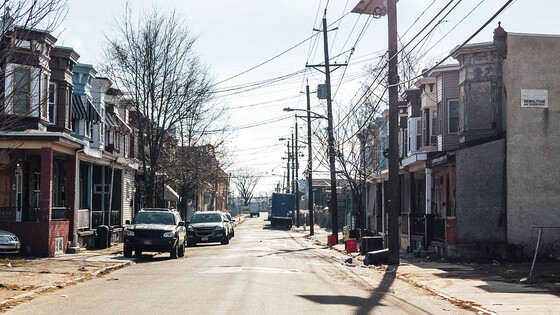 Cars and trucks parked on a street with row houses. Electrical wires and street lights are visible above.