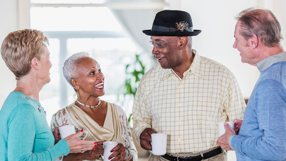 A group of six multi-ethnic seniors standing and talking while holding coffee mugs.