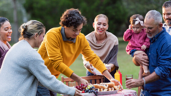 People in a multigenerational family set up a picnic table with plates of hot dogs and fruit.