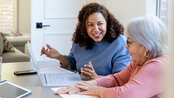 An older woman speaks to a younger woman who is taking notes by hand and on a laptop computer.