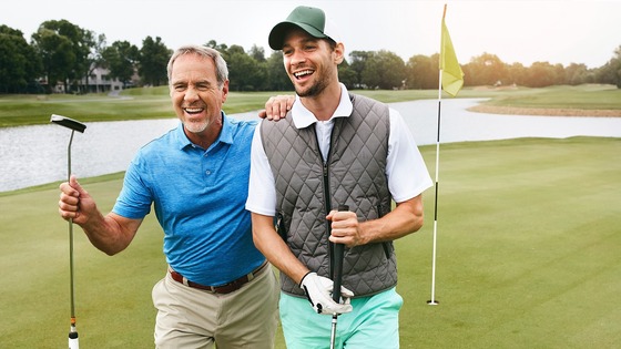 Two men holding putters stand on a golf course green.