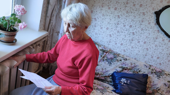 An elder woman sits by her window while reading a packet of papers.