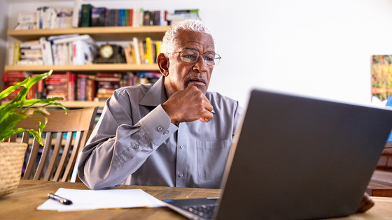 An African American man works at home on his laptop. Bookshelves are in the background.