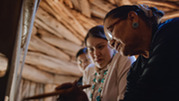 A grandmother teaches her teenaged granddaughters how to weave on a loom.