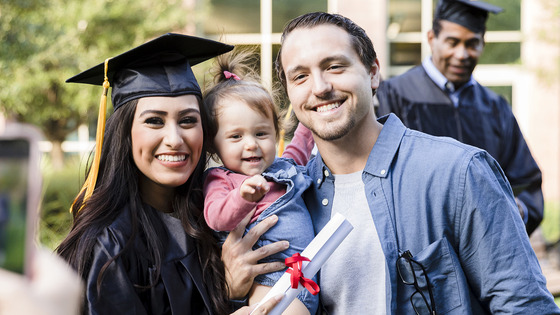 A graduate in a cap and gown poses outdoors with another person and a baby. 