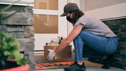 A masked and gloved person crouches on a front porch to set down a box of groceries near the door.