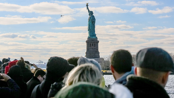 A crowd of people looks toward the Statue of Liberty.