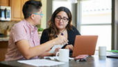 Two people sit at a kitchen table reviewing paperwork while looking at a laptop. 