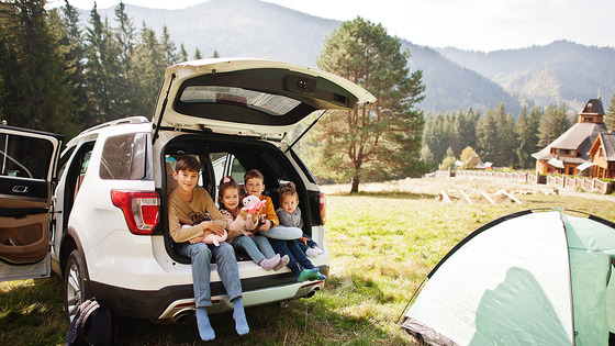 A group sits in the open trunk of an SUV at a campsite.
