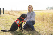 Woman wearing USDA shirt kneeling next to a black working dog wearing a USDA jacket.