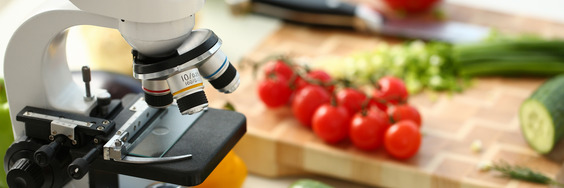 A laboratory microscope next to ripe tomatoes, cucumbers, and a wooden cutting board