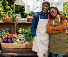 Man and woman stand in front of their produce stand at a farmer's market. Image courtesy of Getty Images.