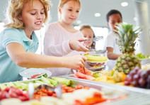 Children choose fruits and vegetables for lunch in a cafeteria, courtesy of Adobe Stock.