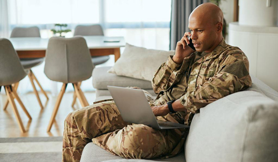 Military veteran on couch looking at computer, courtesy of Adobe Stock.