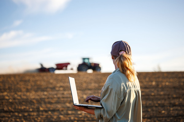 Woman farmer with laptop analyzing agricultural data in front of tractor and seeding machine on cultivated field