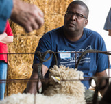 A black man seated w/lamb in front of him and haybales behind him