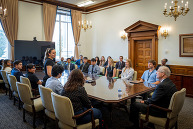 A group of about 20 college students and adults sits around a conference table. Once student stands to speak.