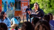 A woman standing and speaking to a seated group in an outdoor setting