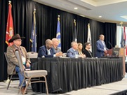 A group of veterans seated on a stage; one standing at the podium speaking