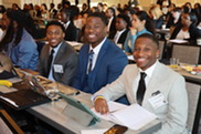 3 young Black men seated with wide smiles seated at front of a full lecture hall