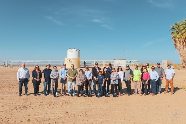 A group gathers at a groundbreaking event, holding shovels in dirt with water tanks behind them.