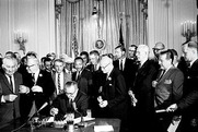 President Lyndon B. Johnson sits at a desk and signs the Civil Rights Act of 1964 among several notable onlookers.