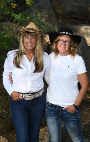 Two people stand outside next to each other while smiling. They are both wearing white shirts, cowboy hats, and jeans.