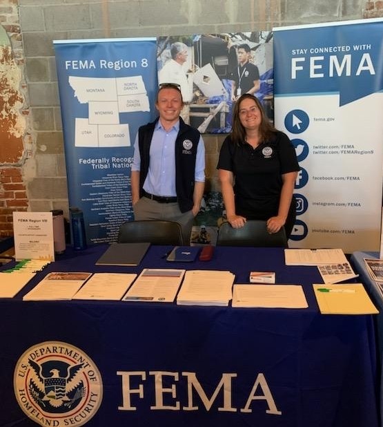 Two FEMA employees stand behind a table with a FEMA tablecloth on it.