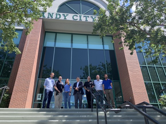 Seven people pose facing the camera at the top of a flight of stairs in front of a building with large glass windows and brick columns.