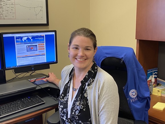 Katherine Fox sits at a desk with a computer screen displaying the Great ShakeOut website.