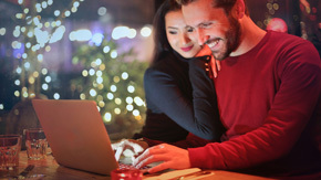 2023 Nov M3 A happy couple seated together looking at a computer with Christmas lights in the background