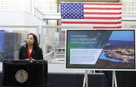 Assistant Secretary Katie Jereza at a podium in front of the American flag and next to a poster titled "Microgrid Collaborations: From Lab to Grid"