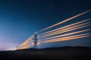 A silhouette of a transmission tower under a starry night sky, with power lines glowing as bright, diagonal orange light trails.