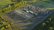 Aerial view of an electrical substation with complex steel frameworks, transformers, and power lines, surrounded by green fields and trees.