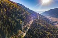 Aerial view of transmission lines cutting through a dense mountain forest, with sunlight shining over the valley landscape.