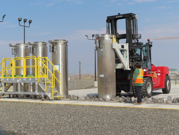 A worker stands on a concrete pad as another worker driving a forklift puts as large column on the pad.  