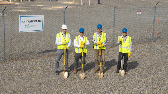 Four people wearing safety vest and hardhats stand together while holding shovels. 