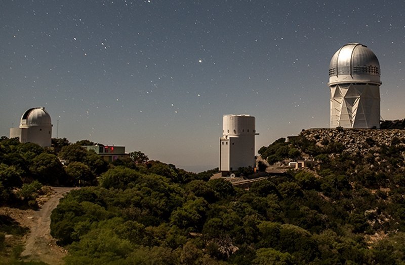 Photo of an observatory with a domed ceiling and a few other white buildings on a hill with the night sky in the background