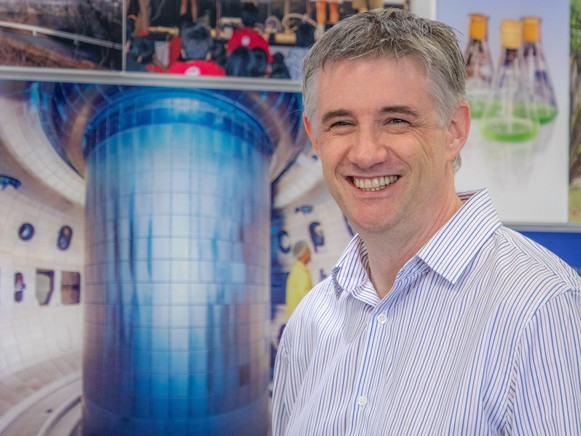 Photo of a white man in a button-down shirt in front of a poster of a tokamak