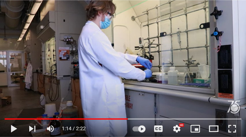 Screenshot of a video showing a person in a mask and lab coat working in a chemistry workspace in a lab
