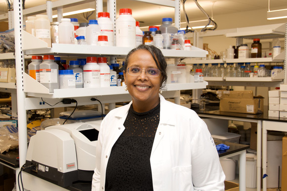 Photo of Dr. Asmeret Berhe, a Black woman in a lab coat standing in a lab full of chemical supplies