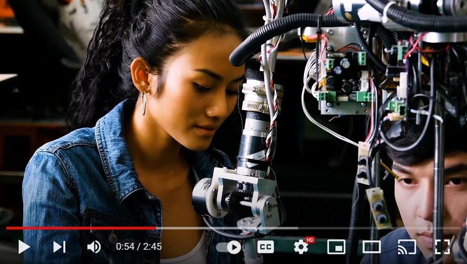 Screenshot of a video that shows a Southeast Asian woman and an East Asian man working on a complex piece of scientific equipment