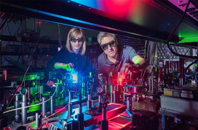 Photo of a white woman and man working with a tabletop of scientific equipment, including lasers and optics equipment