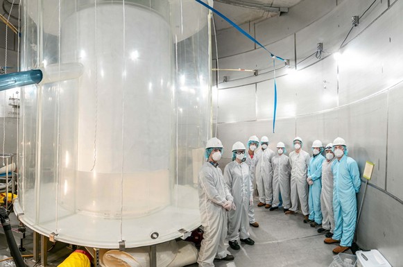 Photo of a group of people in clean room suits and masks standing next to a large clear cylinder with a white cylinder inside it