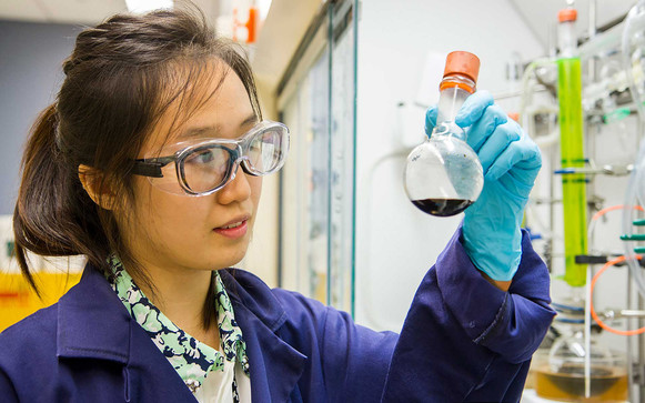 Photo of an Asian woman in lab goggles and gloves holding up a flask with a dark liquid in it