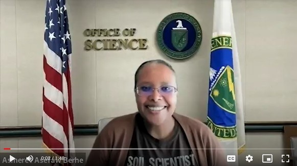 Screenshot of a video of Asmeret Asefaw Berhe looking at the camera and smiling with the American and DOE flags behind her